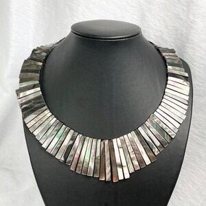 Könnte beinhalten: Eine silberne und schwarze, irisierende Halskette aus rechteckigen Muschelstücken. Die Halskette wird auf einer schwarzen Mannequin-Büste präsentiert.