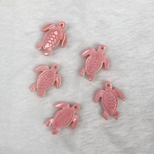 Può includere: Cinque ciondoli a forma di tartaruga marina rosa con intagli dettagliati. I ciondoli sono realizzati in un materiale liscio e lucido e hanno un'estetica delicata e femminile.