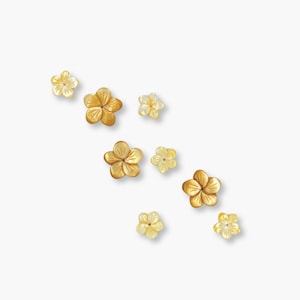 May include: A collection of small, flower-shaped jewellery pieces in shades of gold and yellow. The floral designs are arranged diagonally against a white backdrop, highlighting their delicate details and textures. These pieces appear to be earrings or similar accessories.