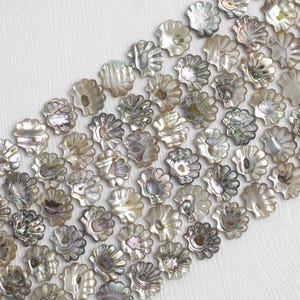 Pode incluir: Close-up de contas iridescentes em forma de concha, em tons de branco, prata e cinza. As contas são dispostas diagonalmente sobre um fundo branco, mostrando suas superfícies texturizadas únicas.
