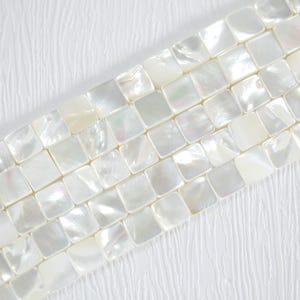 Może przedstawiać: Zbliżenie na sznur kwadratowych, opalizujących pereł z masy perłowej. Koraliki mają kremowo-biały kolor i subtelny połysk. Koraliki ułożone są po przekątnej na białym, teksturowanym tle.