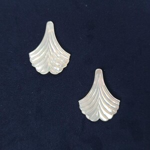 Puede incluir: Dos colgantes de concha de color blanco roto iridiscente con forma de abanicos estilizados. Cada pieza presenta una superficie lisa y curva con crestas verticales que irradian desde un punto superior estrecho hacia un borde inferior ancho.