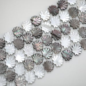 Pode incluir: Close-up de contas em forma de concha em tons de branco, cinza e preto iridescente. As contas estão dispostas diagonalmente, mostrando sua estrutura detalhada em forma de leque. Ideais para a criação de joias e projetos de artesanato.