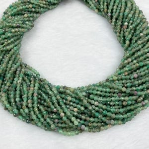 Può includere: Fili di piccole perline verdi sfaccettate. Le perline sono un mix di verde chiaro e scuro con alcuni accenti rosa-rossastri. Le perline sono strettamente infilate insieme, formando una massa densa e strutturata. Le perline sono probabilmente per la creazione di gioielli.