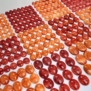 May include: A collection of oval-shaped, polished stones in various shades of red and orange. The stones are arranged in rows on a white surface.