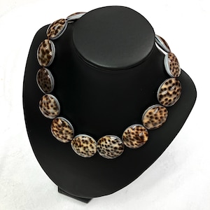 May include: A necklace featuring large, oval-shaped beads with a speckled brown and tan pattern. The beads are strung together, creating a statement piece. The necklace is displayed on a black jewelry stand.