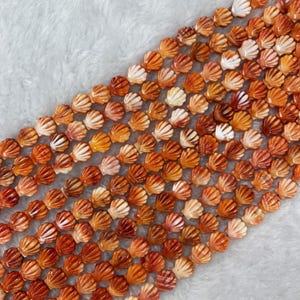 Pode incluir: Close-up de fios de contas de concha laranja e branca. As contas são esculpidas em uma forma detalhada de concha, com uma mistura de tons laranja e branco. As contas são enfiadas juntas, criando um padrão texturizado e colorido.