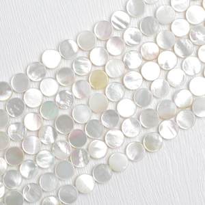 Puede incluir: Primer plano de una colección de cuentas redondas de nácar iridiscente. Las cuentas están dispuestas en filas diagonales sobre un fondo blanco texturizado. Las cuentas tienen un brillo nacarado, que refleja la luz.