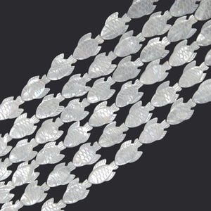 Puede incluir: Filas de pequeñas cuentas iridiscentes con forma de pez sobre un fondo negro. Las cuentas, con escamas y aletas detalladas, están hechas de un material nacarado. Los peces están dispuestos en diagonal, creando un patrón.