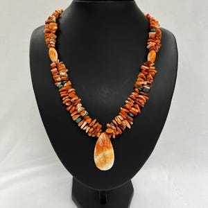 Könnte beinhalten: Eine zweireihige Halskette aus orangefarbenen und weißen Korallenperlen mit einem großen, tropfenförmigen Anhänger. Der Anhänger ist ebenfalls aus Koralle und hat ein weiß-oranges Muster.