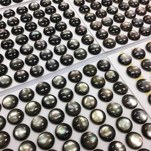 May include: An overhead shot displays numerous dark, lustrous cabochons arranged on white backing cards. The cabochons have a rounded, oval shape and a reflective surface, with hints of iridescent color. The overall impression is of a collection of gemstones.