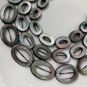 May include: Close-up of a necklace featuring oval-shaped, iridescent beads. The beads are a mix of dark gray, silver, and hints of green and purple. Each bead is strung with a black thread, creating a layered design.