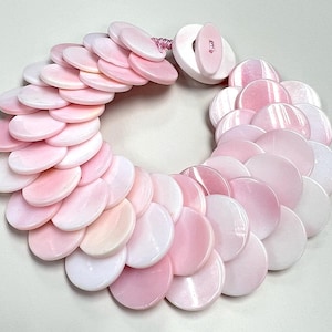 May include: A pink and white necklace composed of numerous flat, round, shell-like pieces. The necklace features a gradient of colors, transitioning from a deep pink to a soft white. The clasp is a large, round, white button.