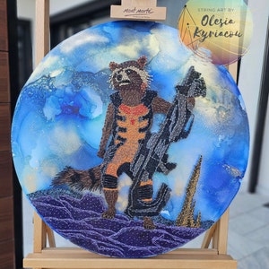 May include: String art of Rocket Raccoon from Guardians of the Galaxy, standing on a purple and blue background. The art is created with string and nails, and the background is painted with blue and purple ink.