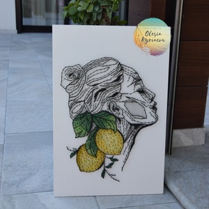 Op de afbeelding: Een zwart-wit portret van een vrouwenhoofd met citroenen en bladeren, gemaakt met draad en spijkers. De kunst is op een wit canvas met een kleine ronde sticker in de hoek die "String Art by Olesia Kypriakov" zegt.