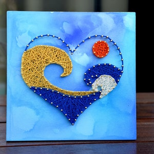May include: A square canvas art piece featuring a heart-shaped design made with string. The heart is divided into sections of gold, blue, and white string, with a small orange circle at the top. The background is a light blue watercolor wash.