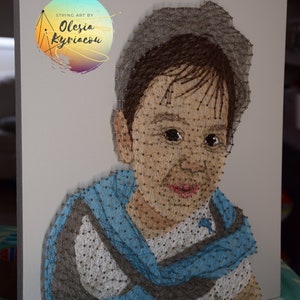 Pode incluir: Retrato de uma criança com cabelo castanho usando uma camisa azul e um chapéu cinza, feito com arte de barbante. A obra é criada com pregos e barbante em uma tela branca. O nome da artista, "Olesia Kyriacon", está escrito em um círculo no canto superior esquerdo da tela.