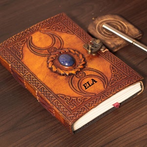 Handmade Brown Genuine leather bound journal Book of shadows grimoire featuring Triple Moon emboss, secured with swing clasp closure, showing blue stone ideal as spell book, travel dairy, rustic guestbook, rest on vintage table with a pen.