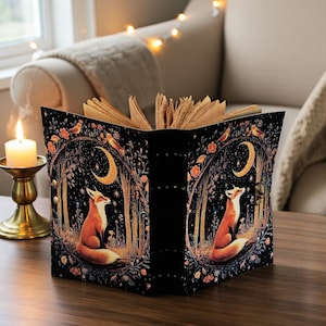 Può includere: Un libro aperto con una copertina nera raffigurante una scena boschiva fantastica con una volpe, una luna e stelle. Le pagine del libro sono invecchiate e una candela accesa è posizionata accanto su un candelabro dorato.