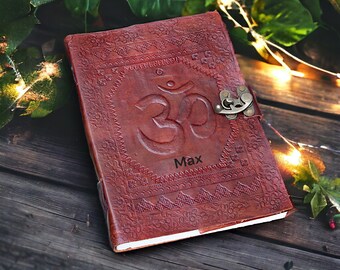 Könnte beinhalten: Ein braunes Lederjournal mit einem geprägten Om-Symbol und dem Namen "Max" auf der Vorderseite. Das Journal hat einen dekorativen Verschluss und ist geschlossen.
