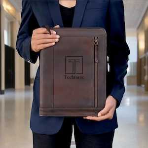 May include: A brown leather portfolio with a zip and the word "Techtonic" printed on the front. The portfolio is held by a person wearing a navy blue blazer.
