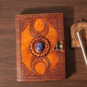 Handmade Brown Genuine leather bound journal Book of shadows grimoire featuring Triple Moon emboss, secured with swing clasp closure, showing blue stone ideal as spell book, travel dairy, rustic guestbook, rest on vintage table with a pen.