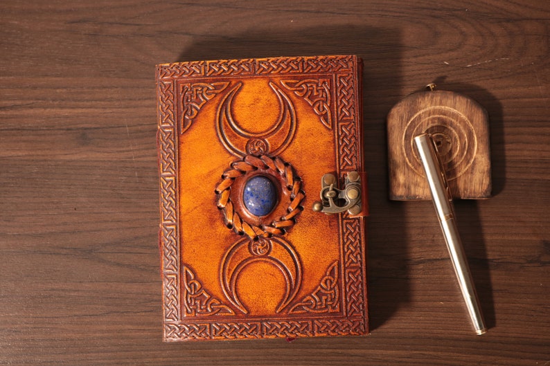 Handmade Brown Genuine leather bound journal Book of shadows grimoire featuring Triple Moon emboss, secured with swing clasp closure, showing blue stone ideal as spell book, travel dairy, rustic guestbook, rest on vintage table with a pen.