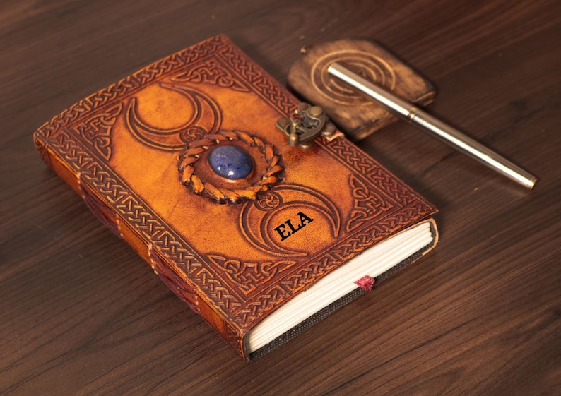 Handmade Brown Genuine leather bound journal Book of shadows grimoire featuring Triple Moon emboss, secured with swing clasp closure, showing blue stone ideal as spell book, travel dairy, rustic guestbook, rest on vintage table with a pen.