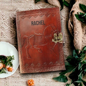 May include: A brown leather journal with a horse design and a brass clasp. The journal has the name "RACHEL" embossed on the front.