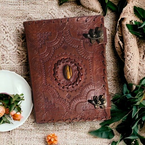 Op de afbeelding: Een bruin lederen dagboek met ingewikkelde gegraveerde ontwerpen en een decoratieve sluiting. Het dagboek heeft een ronde uitsparing met een tijgeroog-steen in het midden.