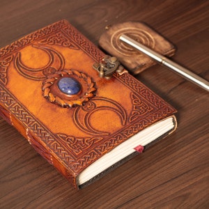 Handmade Brown Genuine leather bound journal Book of shadows grimoire featuring Triple Moon emboss, secured with swing clasp closure, showing blue stone ideal as spell book, travel dairy, rustic guestbook, rest on vintage table with a pen besides.