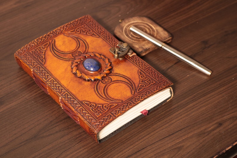 Handmade Brown Genuine leather bound journal Book of shadows grimoire featuring Triple Moon emboss, secured with swing clasp closure, showing blue stone ideal as spell book, travel dairy, rustic guestbook, rest on vintage table with a pen besides.