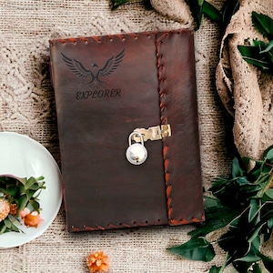 Op de afbeelding: Een bruin lederen dagboek met een slot en sleutel. Het dagboek heeft een gestikt ontwerp en het woord "EXPLORER" is gegraveerd op de voorzijde. Het dagboek ligt op een getextureerd oppervlak met groene bladeren.