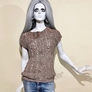 Op de afbeelding: Een bruine top met open weefsel, korte mouwen en een vierkante halslijn. De top heeft een leren accent op de schouder en wordt gecombineerd met een blauwe spijkerbroek. De outfit wordt getoond op een mannequin met grijs haar.