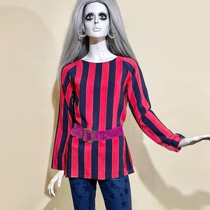 May include: A mannequin wearing a long-sleeved top with vertical red and navy blue stripes, a purple belt, and flared blue jeans with star patterns. The outfit is styled with a vintage aesthetic.