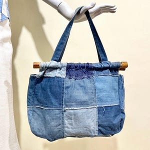 May include: A patchwork denim tote bag with a rectangular shape. The bag features various shades of blue denim squares stitched together. It has denim handles and wooden dowels at the top. The bag is held up by a mannequin hand.