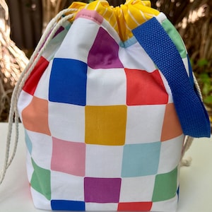 May include: A colourful drawstring bag with a checkered pattern in shades of pink, blue, green, yellow, red, and purple. The bag has a white background and a blue drawstring.