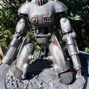 May include: A detailed, metallic robot figurine with a weathered, industrial aesthetic. The robot has a rounded torso, articulated limbs, and a head with red glowing eyes. It stands on a textured, rocky base, showcasing a post-apocalyptic theme.