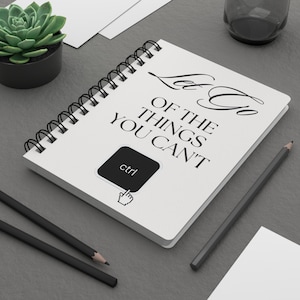 Peut inclure: Un carnet blanc à spirales avec le texte "Let Go Of The Things You Can't" en noir. Un bouton "ctrl" noir avec une icône de main est sur le carnet. Deux crayons noirs et une petite plante succulente en pot noir sont également visibles.