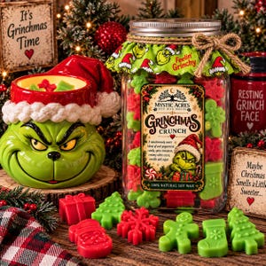 Puede incluir: Una taza de cerámica verde con temática de Grinch y gorro de Santa, junto a un frasco de vidrio con ceras aromáticas rojas y verdes. La etiqueta dice Grinchmas Crunch y Mystic Acres. Ceras sueltas con formas de árboles, calcetines y galletas.