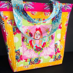 May include: A colourful tote bag with a patchwork design. The bag features a pink, yellow, and green floral pattern with a portrait of a woman on the front. The handles are a light blue with a floral design. The bag is made of quilted fabric.