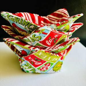 May include: A stack of three fabric bowl cozies with a Christmas theme. The cozies feature red and white striped fabric and a print with the words "Grinch" and "Christmas" in green and red. The cozies are square with folded corners.
