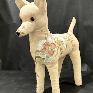 Pode incluir: Uma figura de cão em tecido bege com detalhes florais bordados em tons de branco, pêssego e verde sálvia. O animal de peluche tem olhos de botão castanho-escuro, um nariz preto bordado e orelhas pontiagudas, sobre quatro patas.
