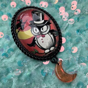 May include: An oval-shaped brooch with a black rope-like border. The brooch features a cartoon owl wearing a top hat and bow tie against a red and yellow background. A small, orange crescent moon charm hangs below the brooch.