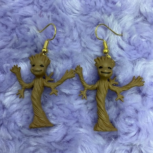 May include: A pair of brown Groot earrings with gold-colored ear wires. The earrings feature a smiling Groot figure with outstretched arms, set against a purple, textured background. The earrings are a fun accessory for fans of the Guardians of the Galaxy.