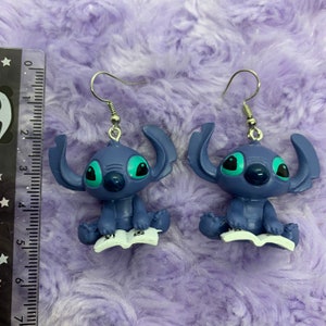 Puede incluir: Un par de pendientes azules de Stitch con ojos verdes. Cada Stitch sostiene un libro y tiene un gancho plateado.