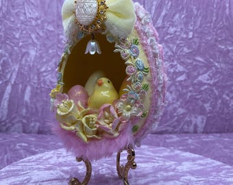 May include: A pink and white Easter egg with a yellow chick inside, decorated with lace, flowers, and a bow. The egg is on a gold stand.