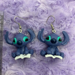 Puede incluir: Un par de pendientes azules de Stitch con ojos verdes. Stitch sostiene un libro en sus manos. Los pendientes son plateados y tienen un cierre de gancho.