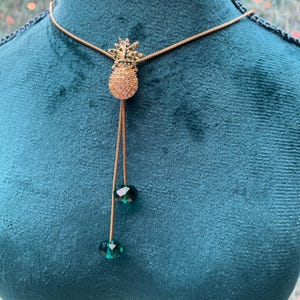 May include: Gold-toned necklace featuring a pineapple pendant adorned with clear and green crystals. The pendant has a textured design and is suspended from a delicate chain. Two green crystal beads dangle from the bottom of the pendant.