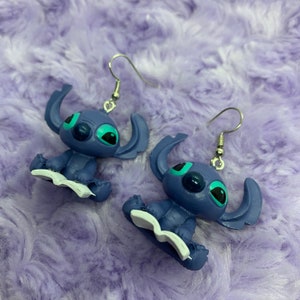 Puede incluir: Un par de pendientes azules de Stitch con ganchos plateados. Cada pendiente presenta a Stitch sentado con un libro en su regazo.
